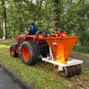 Maschinelle Blumenzwiebelpflanzung. Einoranger Traktor ziet einen Anhänger mit der Pflanzmaschine. Diese besteht aus einem orangen Trichter. Eine Person fährt den Traktor, eine andere Person steht hinter dem Trichter der Pflanzmaschine.