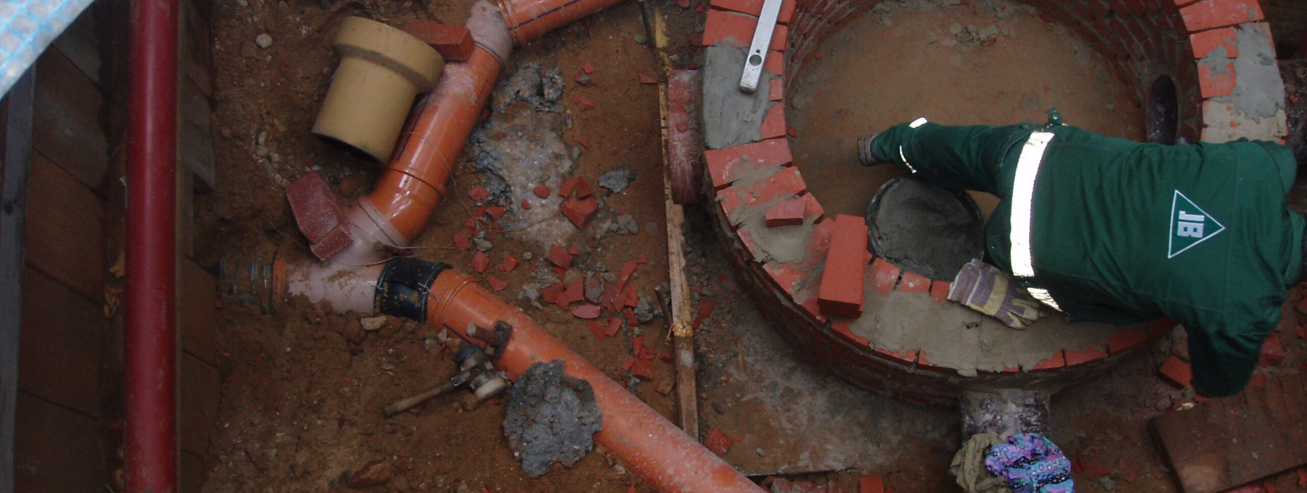 Bauen und Entwässerungsantrag - Baustelle im Fußboden eines Hauses. Zu sehen sind verschiedene Rohr und rote Backsteine. Ein Handwerker mauert einen Schacht.