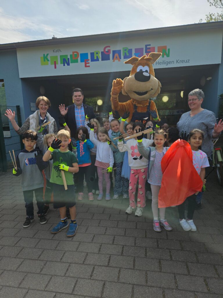 Gruppenbild vor der Kita Zum Heiligen Kreuz. Die Kindergartenkinder zeigen gemeinsam mit erstem Stadtrat Stefan Schmitt und dem DLB Maskottchen Lutz van Putz ihre Müllsammel-Zangen